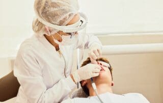 Dentist in surgical uniform, mask, and gloves inserting medical instrument into the patient's mouth during dental treatment.